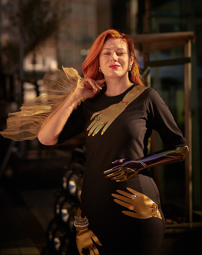 Zdjęcie Aleksandry patrzącej w obiektyw. Metaliczną protezę opiera na biodrze. Prawą ręką dotyka twarzy. Włosy ma spięte złotą spinką nad uchem. Na sukience ma złote aplikacje w kształcie rąk.
