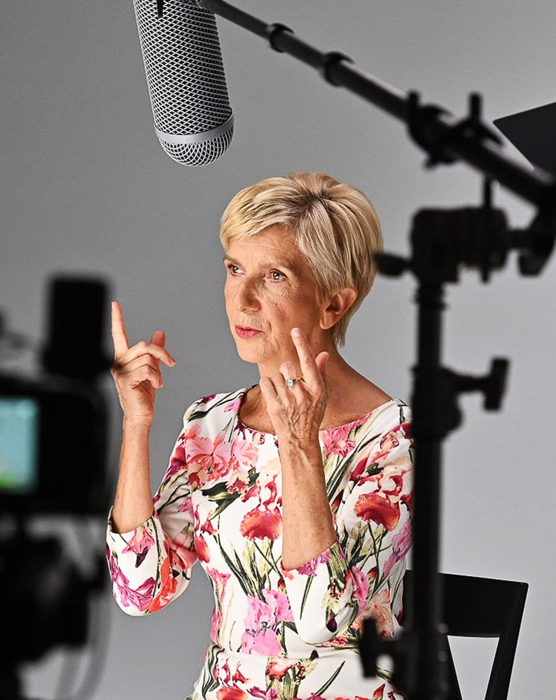 Zdjęcie Barbary z planu zdjęciowego, odpowiadającej na pytanie, gestykulującej. Kobieta ma sobie białą sukienkę z w kwiatowy wzór, siedzi na krześle.