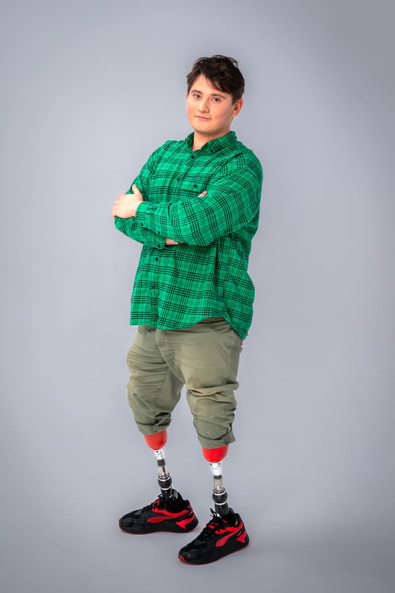 Zdjęcie Bartka stojącego i pozującego na białym tle. Chłopak ma sobie zieloną koszulę w czarną kratę oraz oliwkowe spodnie podwinięte na wysokości kolan odsłaniające dwie protezy nóg w kolorze czerwonym.