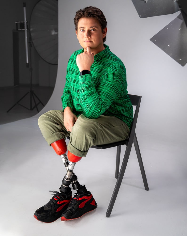 Zdjęcie Bartka z planu filmowego. Chłopak ma sobie zieloną koszulę w czarną kratę oraz oliwkowe spodnie podwinięte na wysokości kolan odsłaniające dwie protezy nóg w kolorze czerwonym. Bartosz siedzi na czarnym krześle ustawionym na białym tle. Patrzy w bok prosto w kamerę i trzyma lewą rękę na brodzie.