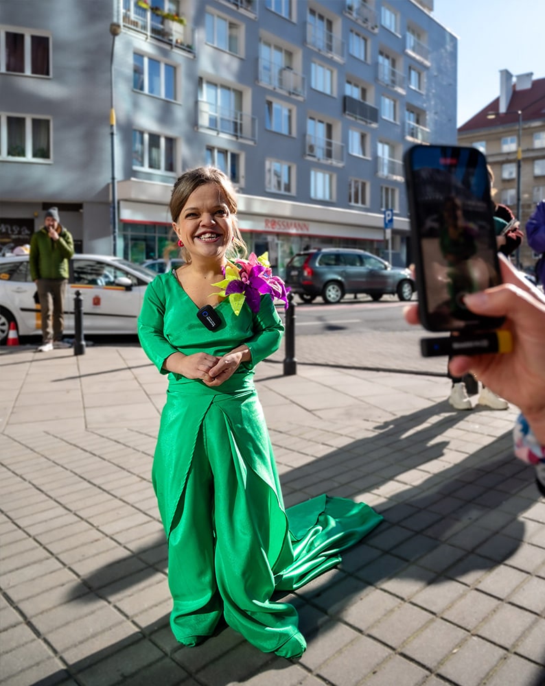 Zdjęcie Ani nagrywanej telefonem i udzielającej wywiadu. Kobieta jest osobą niskorosłą, jest ubrana w elegancką, intensywnie zieloną suknię z trenem i aplikacją w kształcie kwiatów na ramieniu. Przy sukni przypięty ma mikrofon.