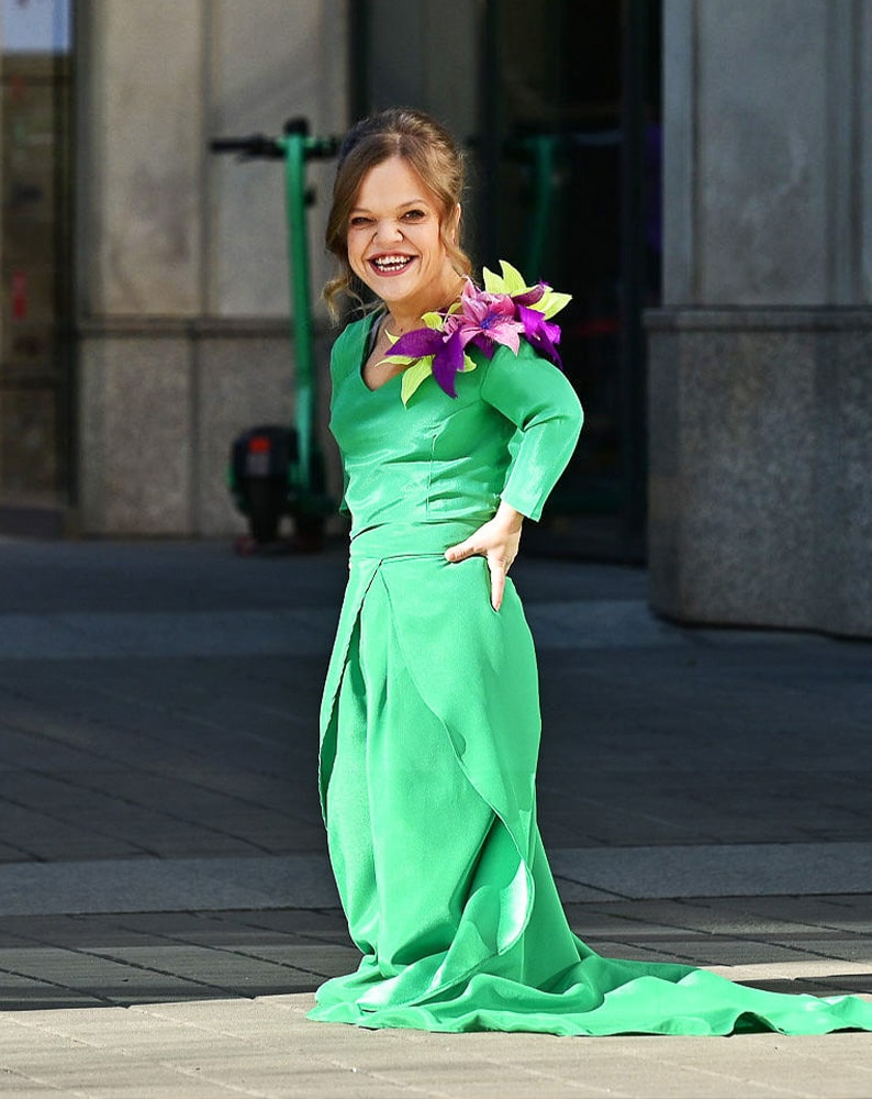 Zdjęcie Ani pozującej z uśmiechem przed obiektywem. Kobieta jest osobą niskorosłą, jest ubrana w elegancką, intensywnie zieloną suknię z trenem i aplikacją w kształcie kwiatów na ramieniu. Jedną rękę ma opartą o biodro.