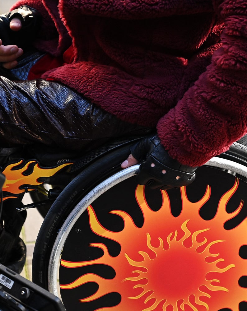 Zdjęcie fragmentu wózka Alana. W bliskim kadrze widać koło z grafiką przedstawiającą żółte płomienie oraz dłoń Alana w skórzanej czarnej rękawiczce dotykającą szprychy koła.