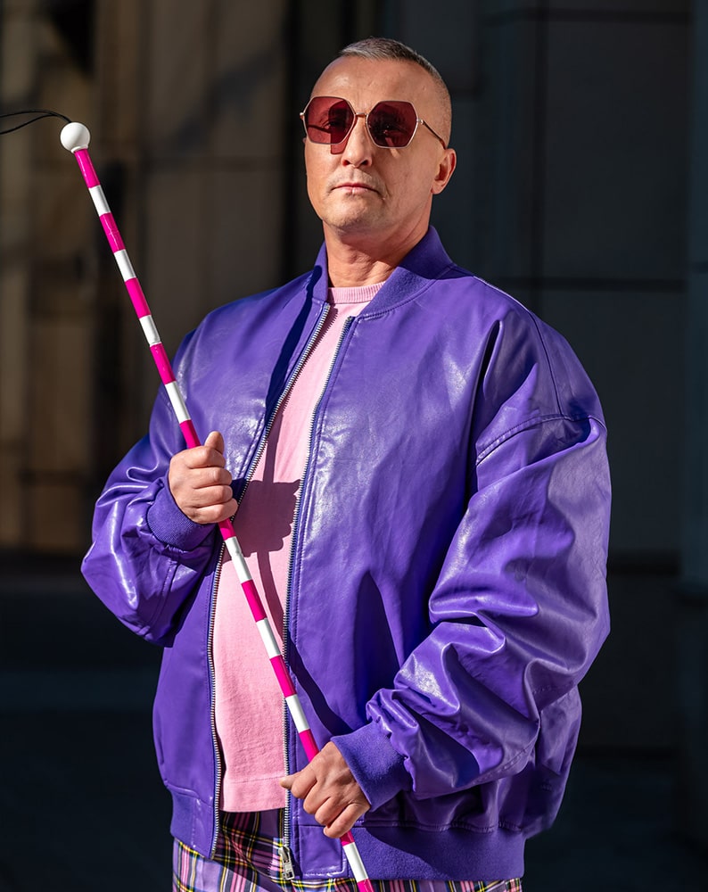 Zdjęcie Sebastiana stojącego na planie zdjęciowym i mówiącego do kamery. Mężczyzna nosi różowe okulary, jest ubrany w intensywnie fioletową skórzaną kurtkę oraz różowy t-shirt. W jednej ręce trzyma laskę dla osoby niewidomej w różowo-białe, ozdobne pasy, drugą ręką drapie się po głowie.