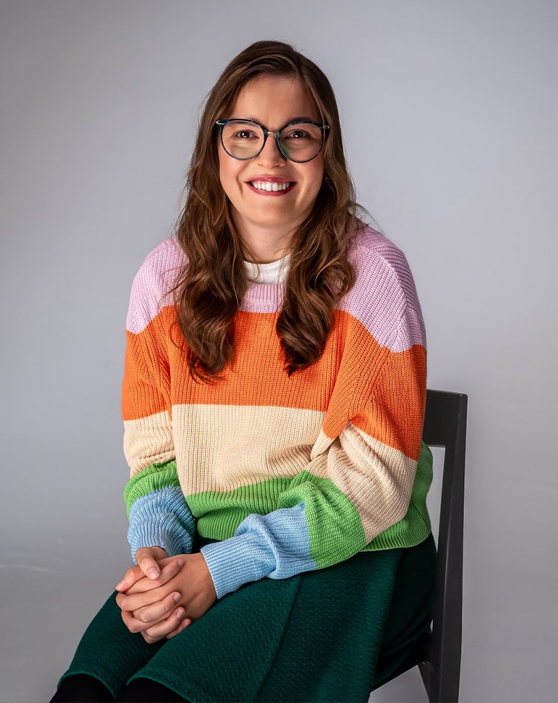 Portret Julii patrzącej w obiektyw z uśmiechem. Dziewczyna ma na sobie sweter w pasy w kolorach: różowym, pomarańczowym, żółtym i zielonym. Nosi okulary korekcyjne.