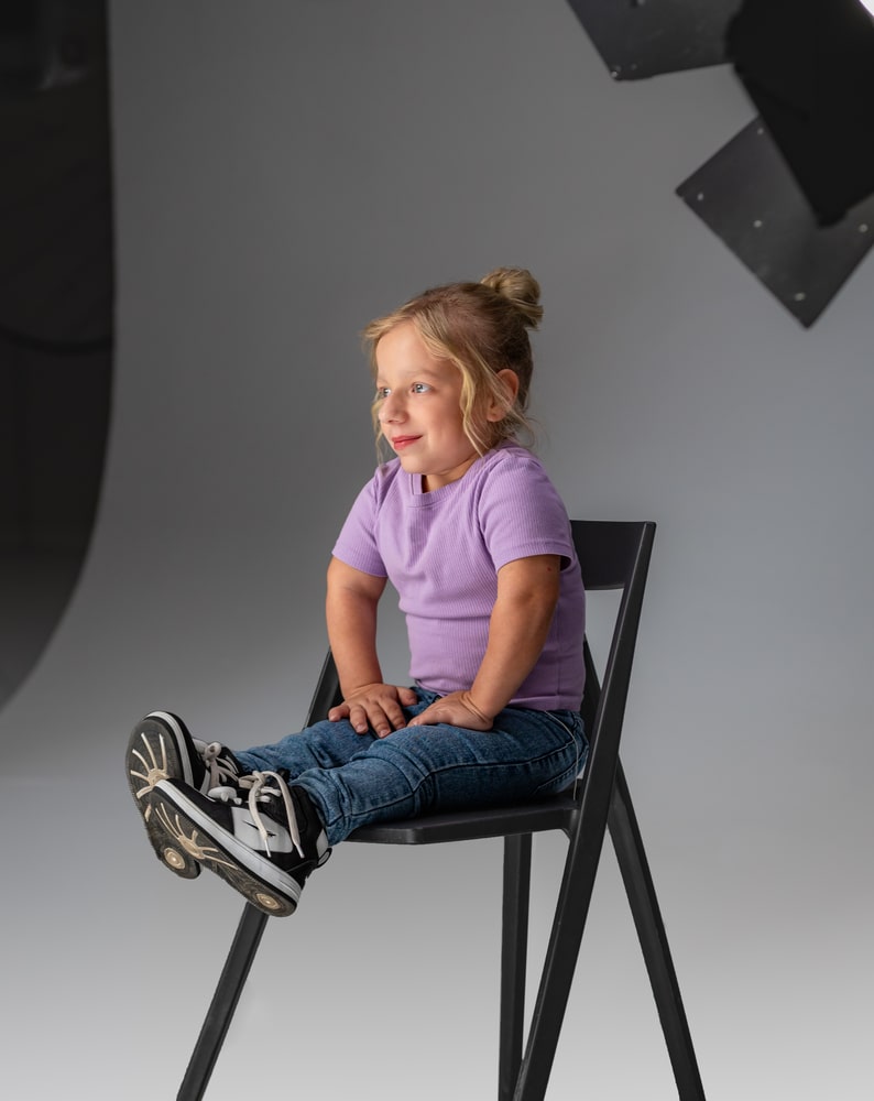 Zdjęcie Victorii z planu filmowego. Dziewczynka jest osobą niskorosłą, ma na sobie koszulkę na krótki rękaw w liliowym kolorze i jeansy, siedzi na czarnym krześle ustawionym na białym tle. Patrzy w bok w kamerę i odpowiada na pytanie.