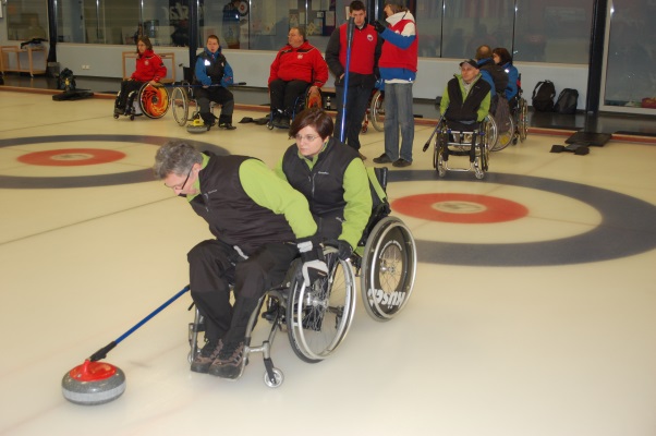 Na zdjęciu para zawodników curlingu na wózkach w trakcie gry. W tle osoby obserwujące rozgrywkę.