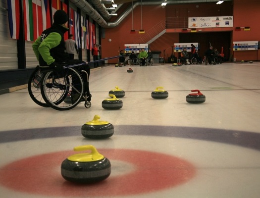 Na zdjęciu zawodnik curlingu na wózku. Na lodzie kamienie w jednej linii.