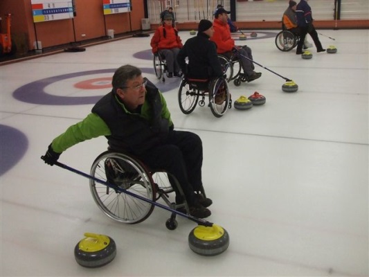 Na zdjęciu zawodnicy curlingu na wózkach. Jeden z nich celuje kamieniem do celu.