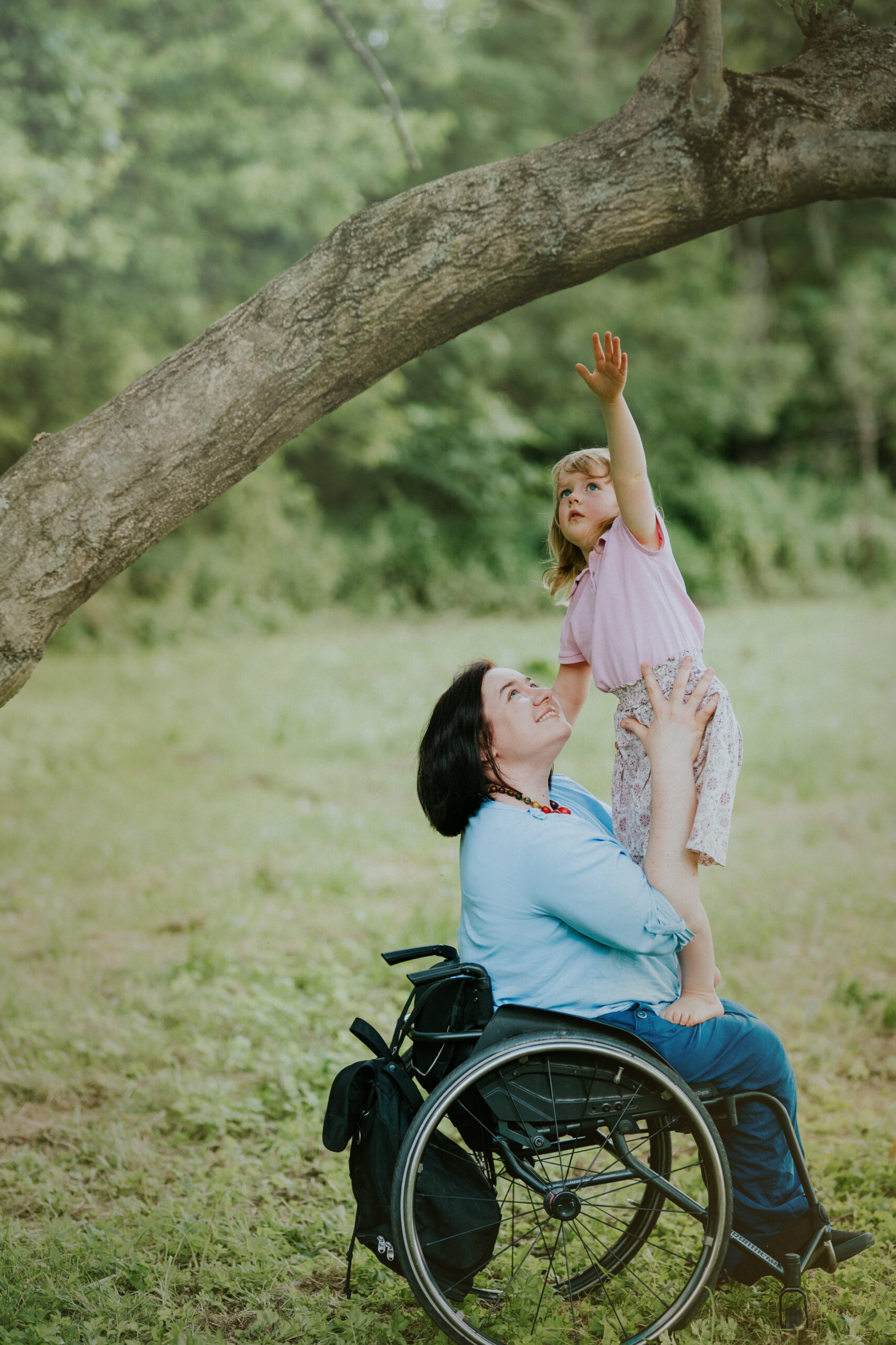 Na zdjęciu Małgorzata Żbikowska na wózku trzyma na kolanach swoją córeczkę. Dziewczynka stoi i sięga do gałęzi drzewa.