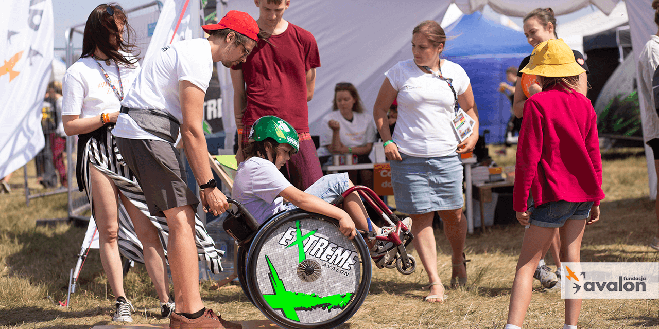  Na zdjęciu chłopiec na wózku pokonuje tor przeszkód z pomocą fizjoterapeuty. W tle namiot fundacji.
