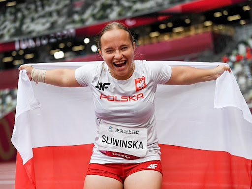 Na zdjęciu znajduje się uśmiechnięta dziewczyna (Renata Śliwińska), która trzyma za plecami dużą flagę Polski. W tle widać trybuny stadionu sportowego.