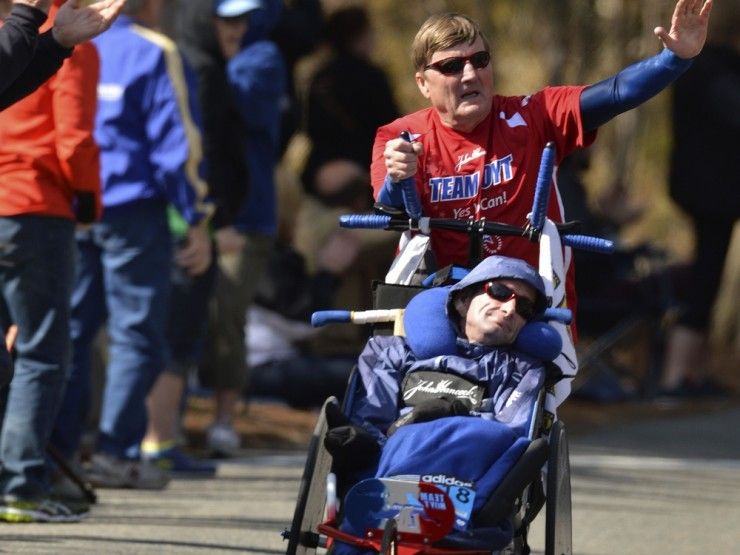 Na zdjęciu starszy mężczyzna w stroju sportowym biegnie, machając ręką i pchając przed sobą młodszego mężczyznę na wózku. 