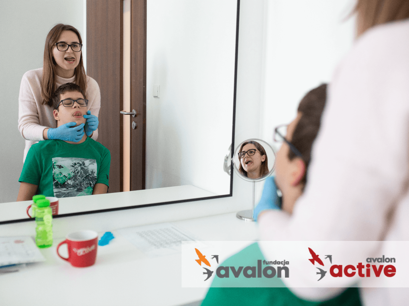 Kobieta i chłopiec stoją przed lustrem i zaglądają mu do buzi. W prawym dolnym rogu zdjęcia znajduje się logo Fundacji Avalon i Projektu Avalon Active.