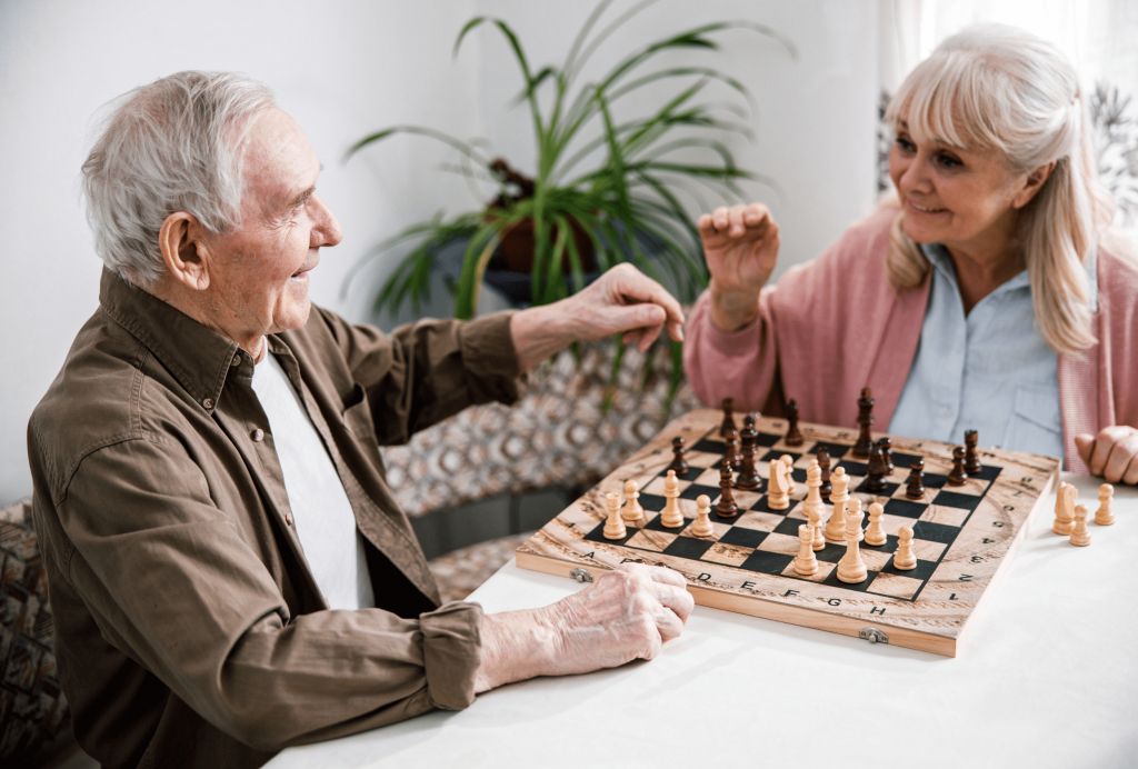 Na zdjęciu znajdują się dwie starsze osoby: mężczyzna i kobieta. Są uśmiechnięci, grają razem w szachy.