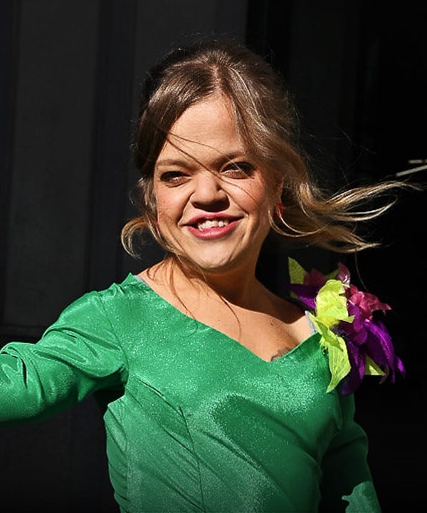 Portret Ani uśmiechające się do obiektywu, z grzywką powiewającą na wietrze. Kobieta jest osobą niskorosłą, jest ubrana w elegancką, intensywnie zieloną suknię z aplikacją w kształcie kwiatów na ramieniu.