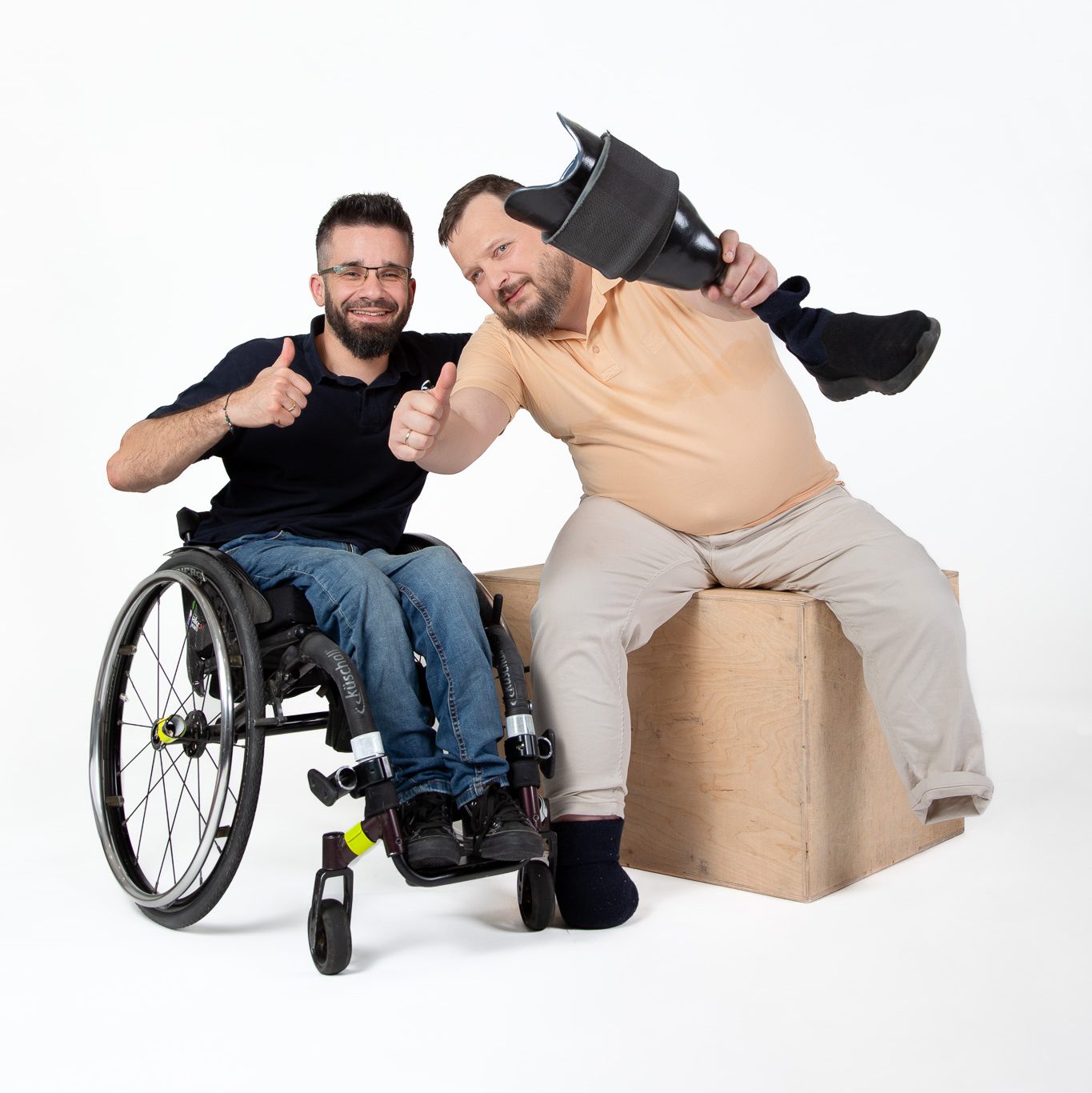 Na zdjęciu dwóch mężczyzn - siedzą przodem do obiektywu, unoszą kciuki w górę i uśmiechają się. Jeden z nich siedzi na wózku, a drugi siedzi na skrzyni do ćwiczeń i trzyma w ręku protezę nogi.