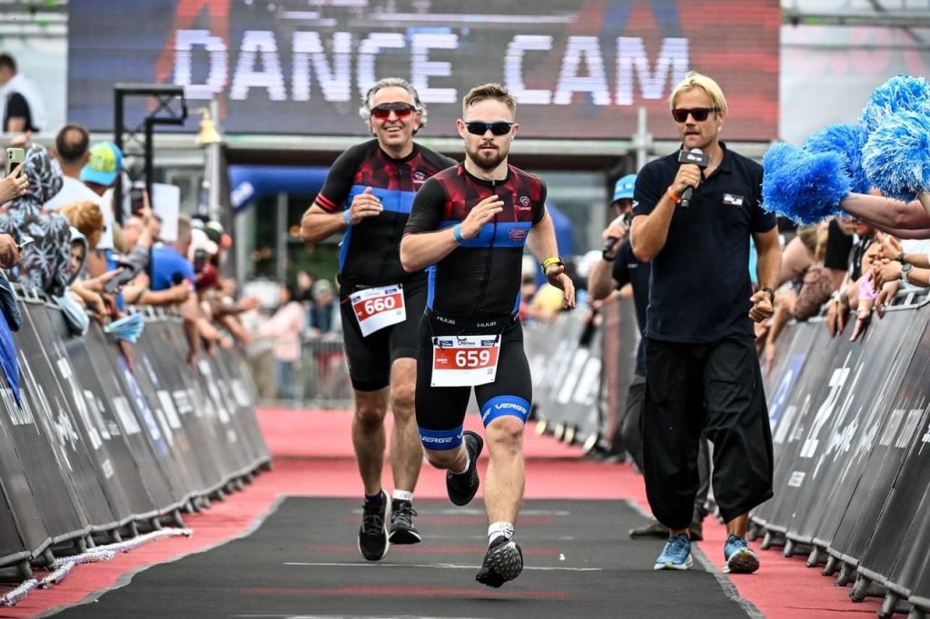 Uśmiechnięty Bartek Matusiewicz biegnie na trasie zawodów, za nim biegnie inny zawodnik, a obok stoi mężczyzna z mikrofonem, który komentuje bieg.