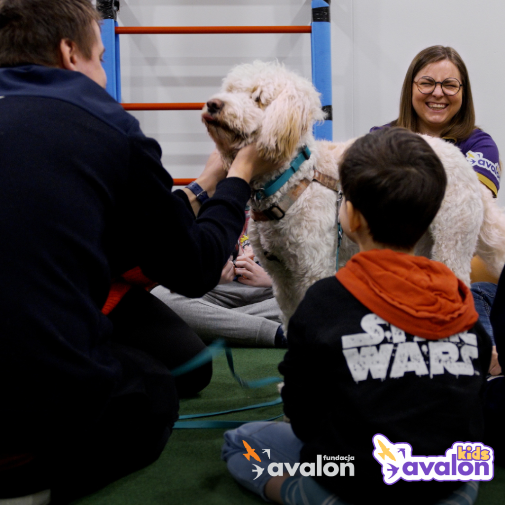 Dzieci głaszczą psa w trakcie dogoterapii, obserwuje ich uśmiechnięta fizjoterapeutka. 