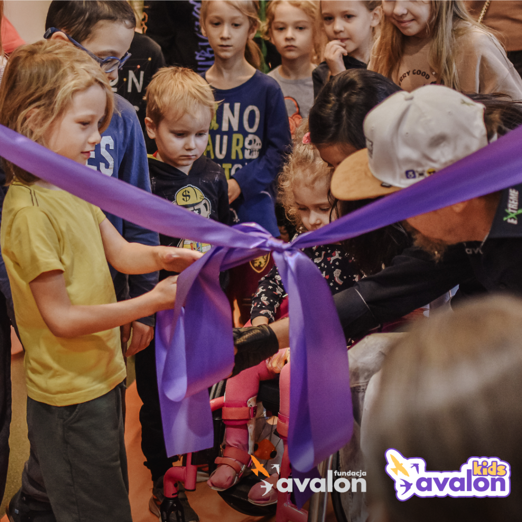 Prezes Fundacji Avalon przecina wstęgę na otwarcie nowej przestrzeni rehabilitacyjnej. Wśród niego znajduje się grupa dzieci, a jeden z małych chłopców mu pomaga. 
