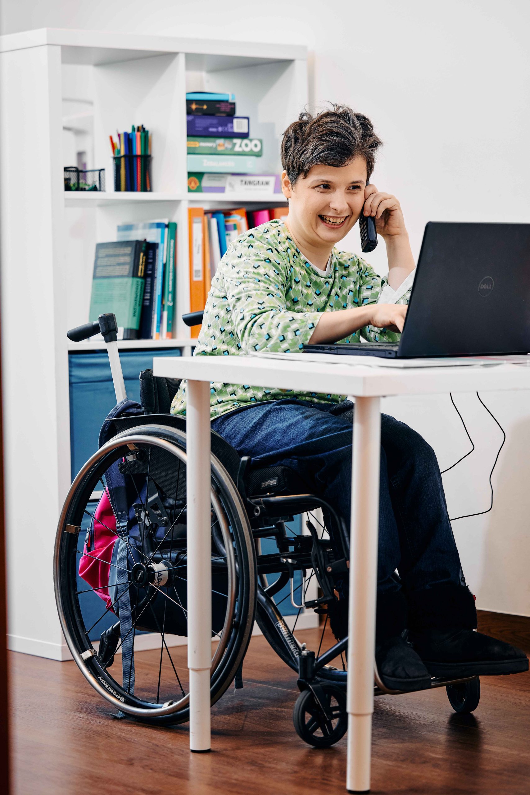 Uśmiechnięta kobieta na wózku siedzi przy komputerze i rozmawia przez telefon