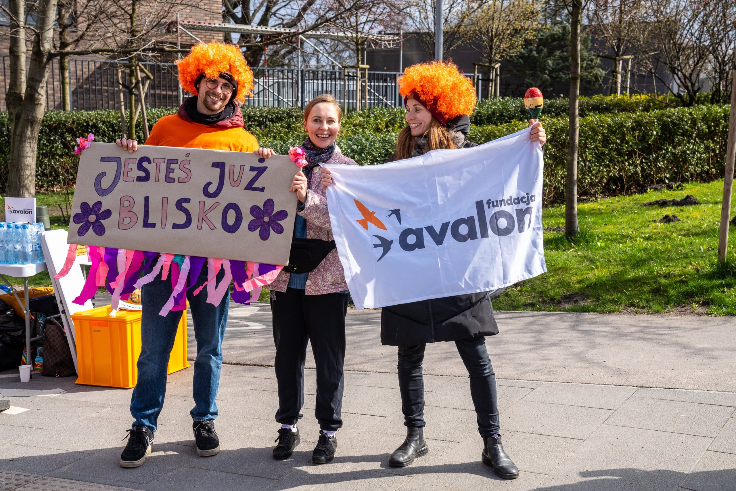 Zdjęcie ze strefy kibica Fundacji Avalon podczas Połmaratonu warszawskiego. Kobieta trzyma flagę z logotypem fundacji Avalon, mężczyzna trzyma transparent z napisem Jesteś już blisko. Oboje mają pomarańczowe peruki. Pomiędzy nimi stoi uśmiechnięta kobieta.