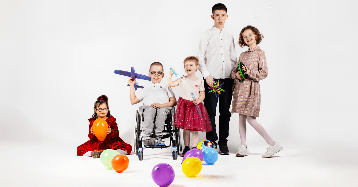 Grupa dzieci z niepełnosprawnościami pełnosprawnych. Wszyscy są uśmiechnięci, pozują do grupowej fotografii wśród kolorowych balonów.