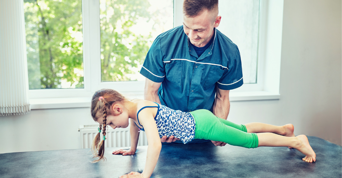 Fizjoterapeuta prowadzi rehabilitację dziecka w jasnym, rozświetlonym dziennym światłem gabinecie. Uśmiecha się i pomaga kilkuletniej dziewczynce utrzymać się w pozycji deski.