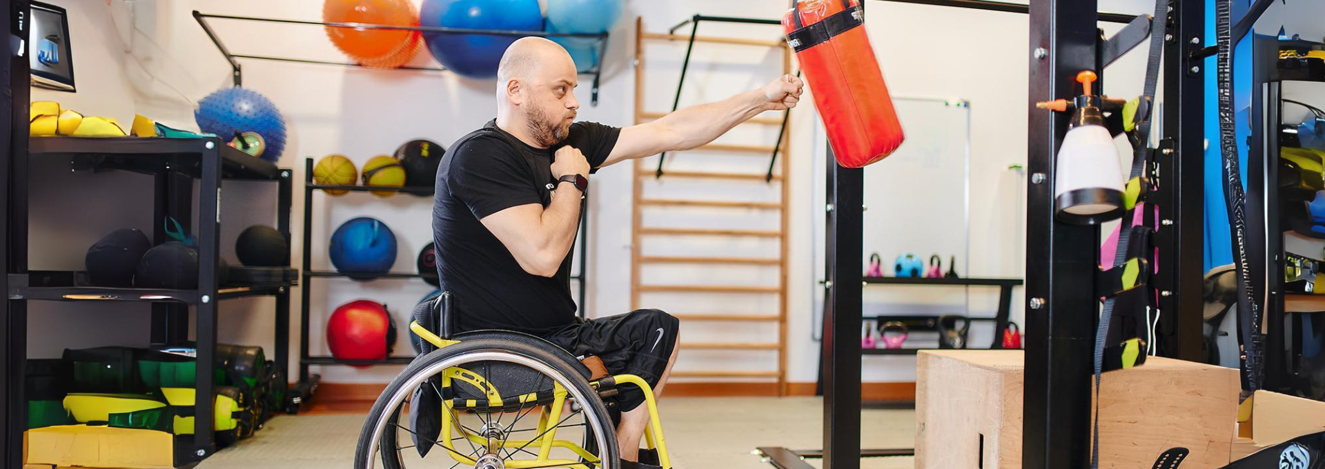 Mężczyzna na wózku boksuje worek treningowy na sali, na której znajdują się różne sprzęty sportowe.