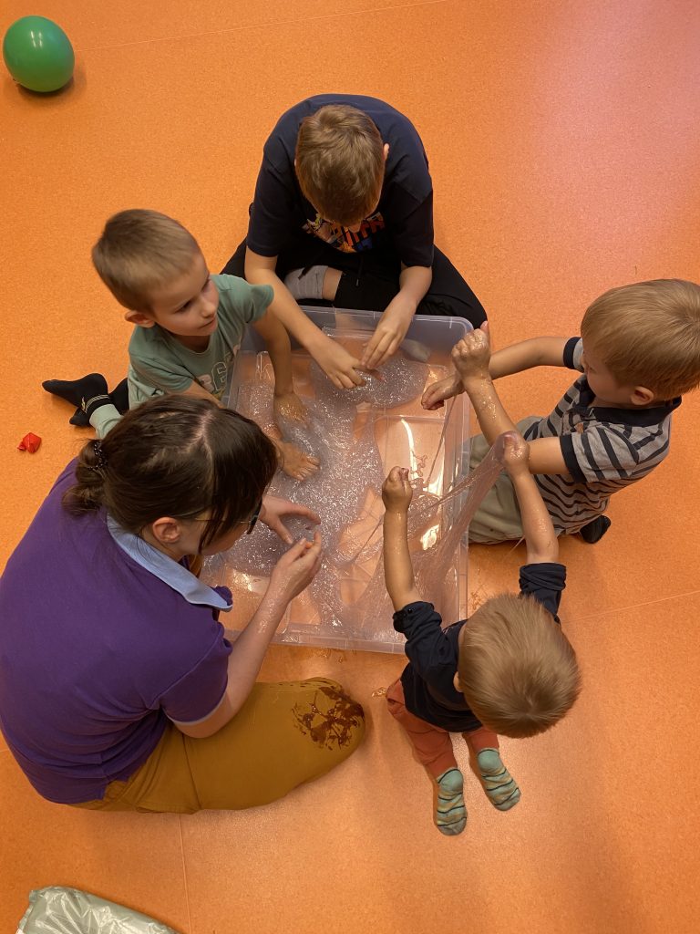 Terapeutka bawi się z czwórką dzieci masą sensoryczną slime. Nachylają się nad pojemnikiem z masą. Zdjęcie jest zrobione z góry. 